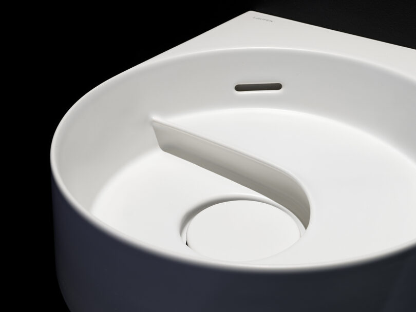 A modern white sink with a unique curved divider and a circular drain, set against a dark background.