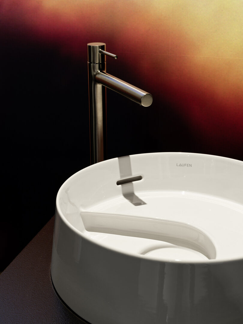 A modern white ceramic sink with a built-in ramp and a tall, metallic faucet, set against a blurred, warm-toned background.