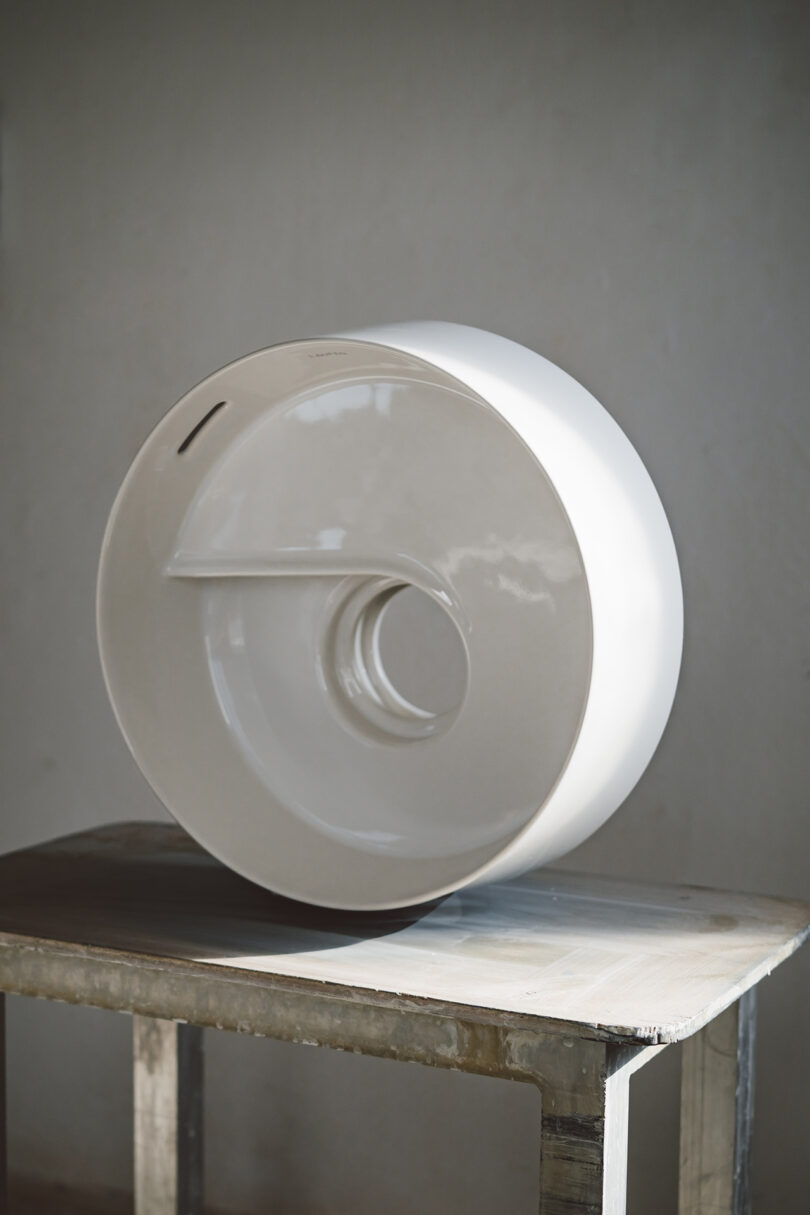 A glossy, white, circular ceramic sink with a central drain hole and a minimalist design, placed on a wooden table against a plain gray wall.