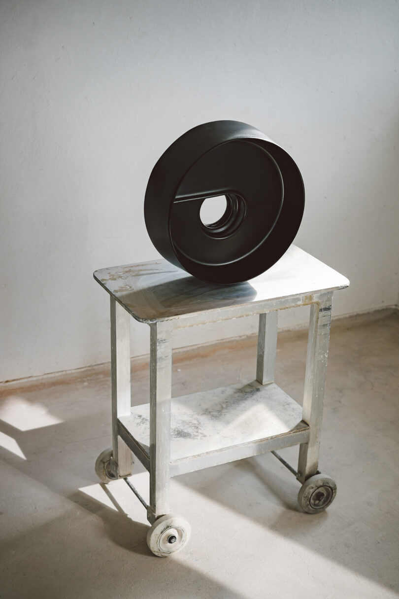 A round, black abstract sculpture with a central hole sits on a small, weathered, white cart with wheels in a bright room.