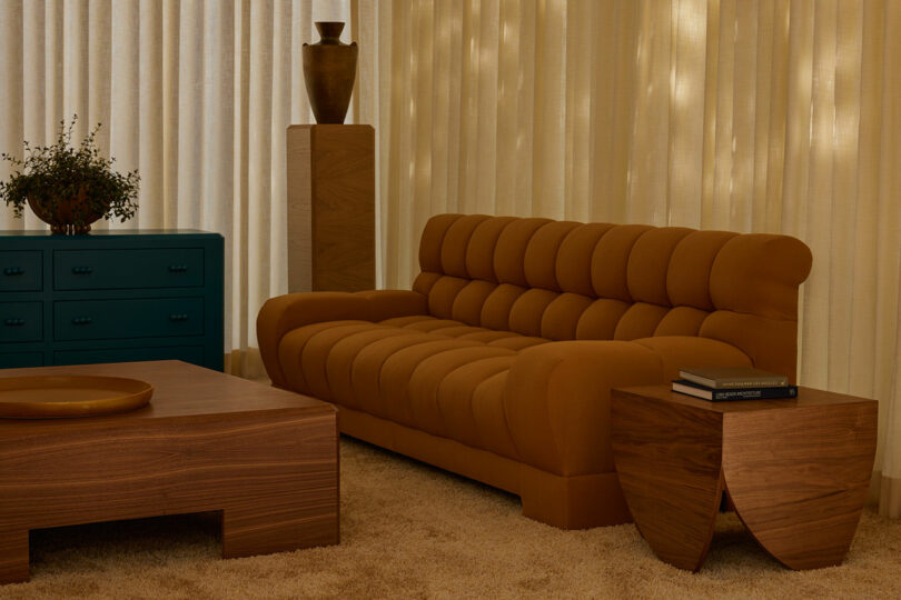 A mid-century modern living room features a tufted mustard sofa, wooden tables, a teal dresser, and floor-to-ceiling curtains, with a vase from the Agapanthus Collection on a pedestal to complete the look.