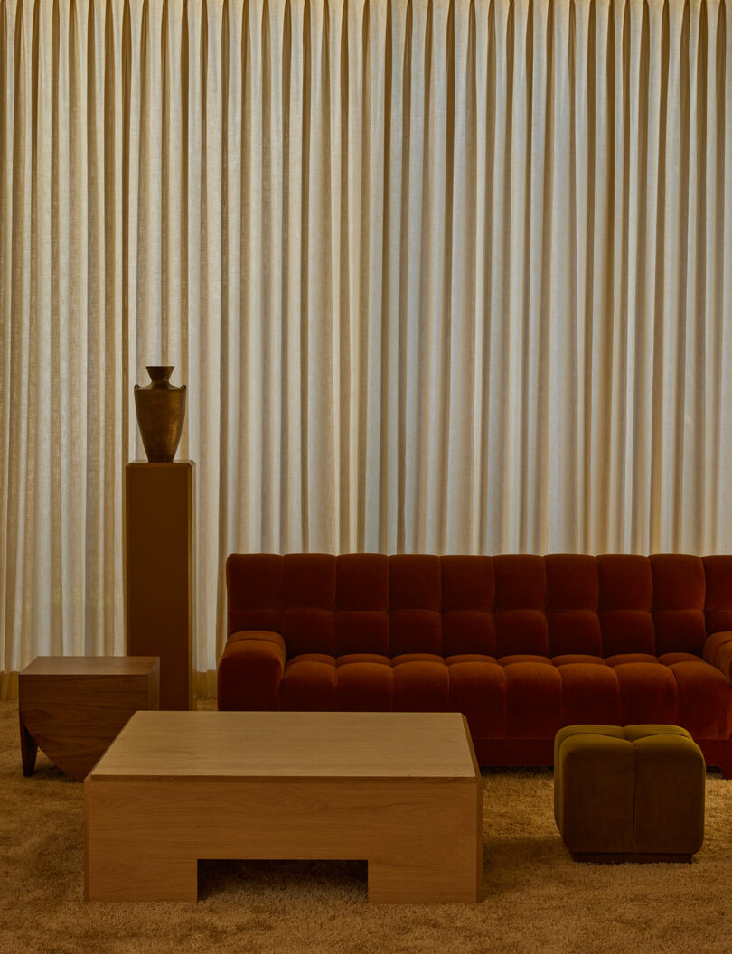 A living room with beige curtains, a rust-colored tufted sofa, a wooden coffee table, side table, brown ottoman, and a ceramic vase from the Agapanthus Collection on a pedestal.