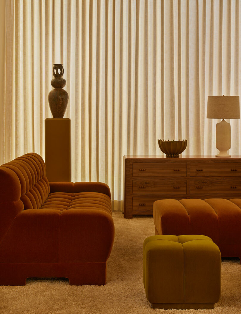 A living room with a tufted rust-colored sofa, matching ottoman, green cube stool, wooden sideboard from the Agapanthus Collection, table lamp, vase, and neutral floor-to-ceiling curtains.