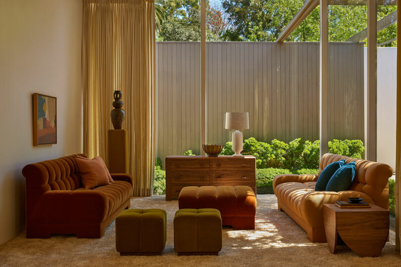A modern living room featuring the Agapanthus Collection: two tufted sofas, an ottoman, two cube stools, a side table, and a wood console set against floor-to-ceiling windows overlooking a lush garden.