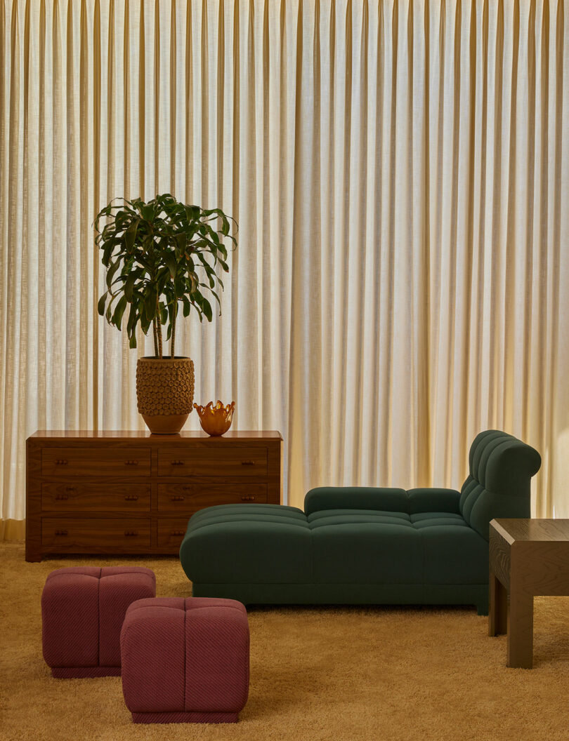 A modern living room featuring a green chaise lounge from the Agapanthus Collection, a wooden sideboard with a potted plant, two red ottomans, a dark side table, and beige carpet and curtains.