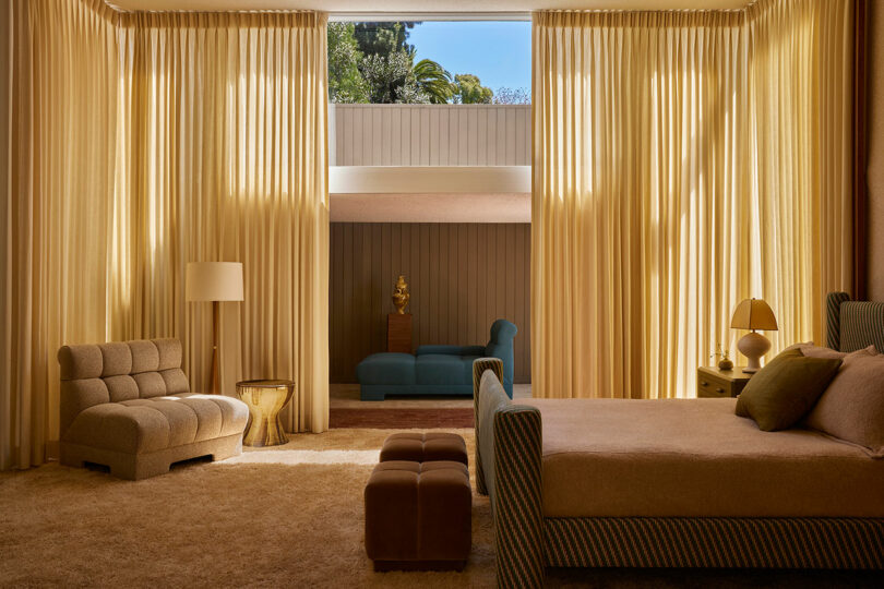 A spacious bedroom with beige curtains, carpet, and furniture from the Agapanthus Collection, featuring a large window, a blue chaise lounge in an adjacent room, and sunlight streaming in.