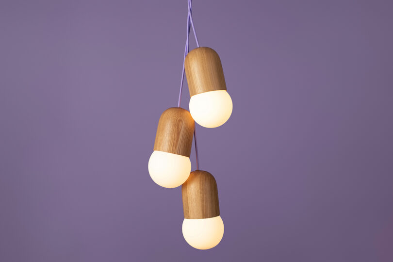 A cluster of three pendant lights with wooden tops and frosted white bulbs hangs against a plain purple background.