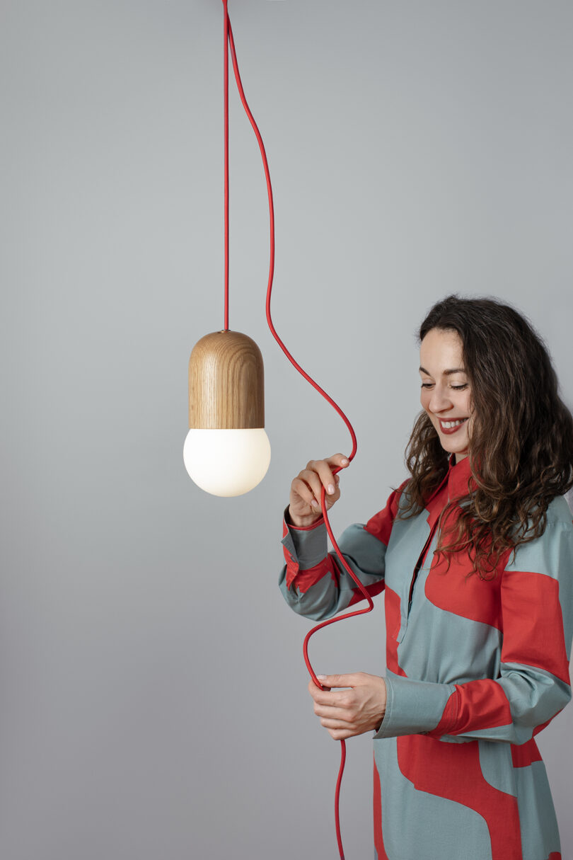 A woman in a patterned dress adjusts a red cord attached to a modern pendant light with a wooden top and glowing bulb, against a plain gray background.