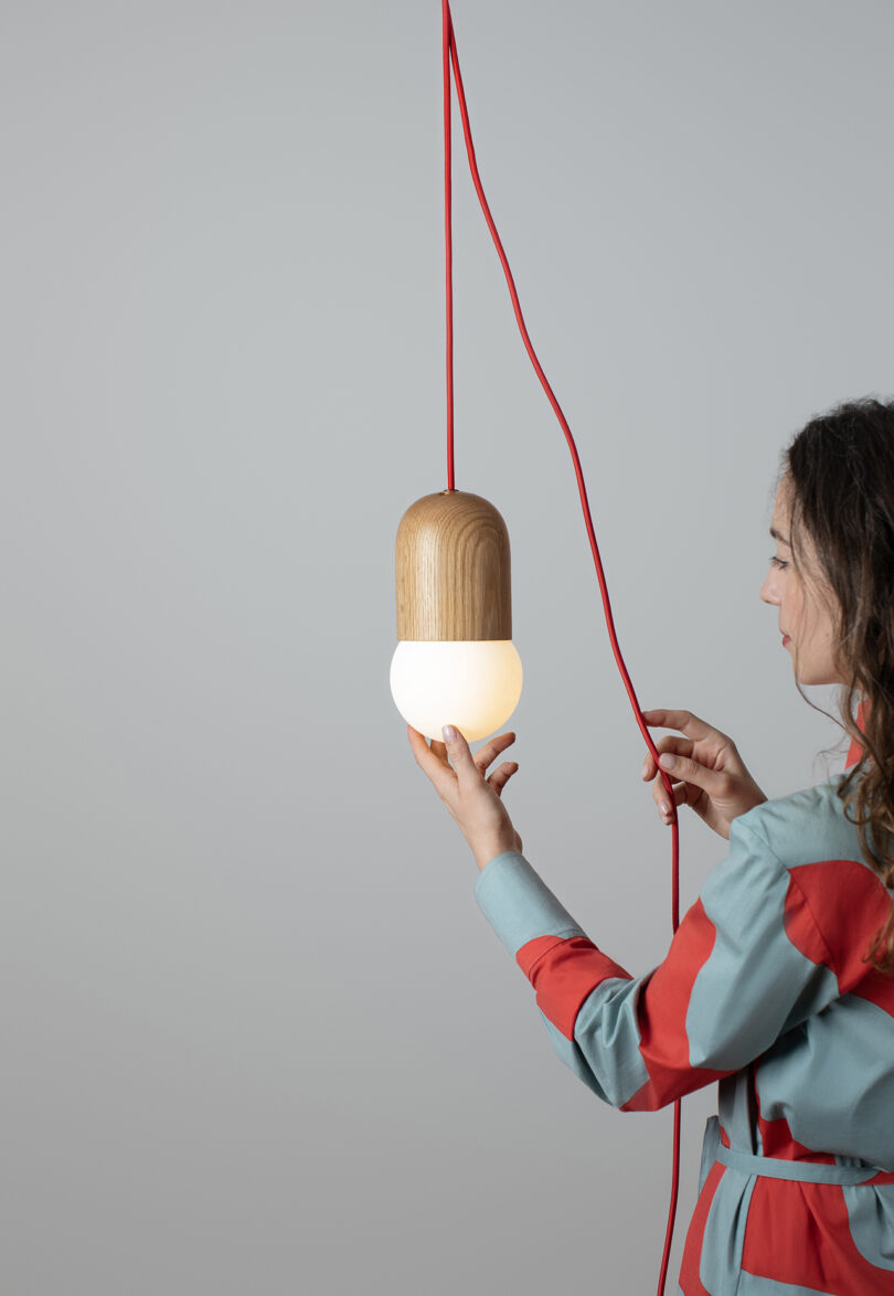A person in a blue and red outfit holds a hanging pendant light with a wooden top and glowing white bottom, suspended by a red cord against a plain background.