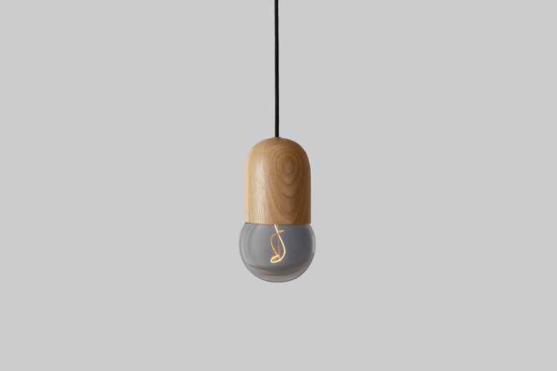 A pendant light with a wooden top and a glass bulb, suspended from a black cord against a plain grey background.