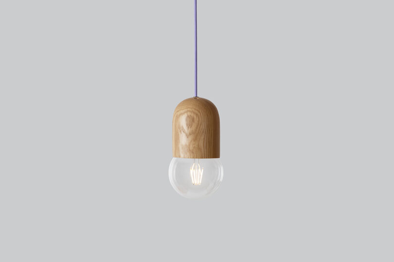 A pendant light with a wooden top and a clear glass bulb hangs from a purple cord against a plain gray background.