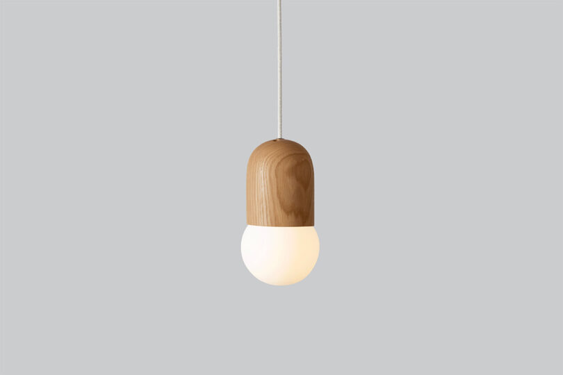 A pendant light with a wooden top and a glowing white bulb hangs from a white cord against a plain grey background.