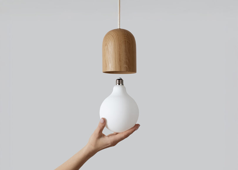 A hand holds a white light bulb below a wooden pendant light fixture against a plain gray background.