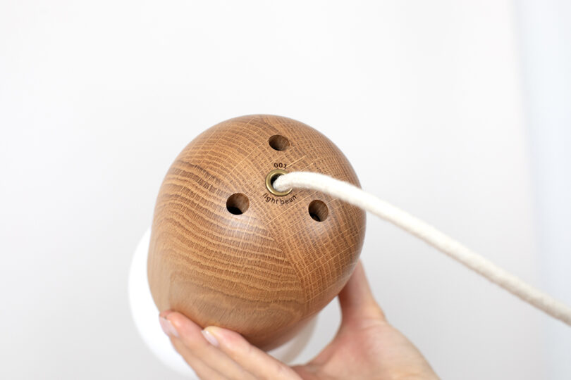 A hand holds a wooden lamp with three holes, and a white rope threaded through a central metal ring.