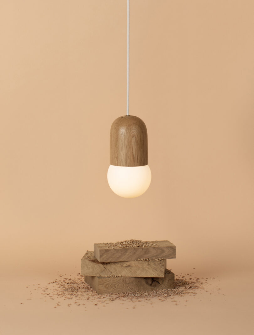 A hanging pendant light with a wooden top and white bulb is suspended above three stacked wood planks and wood shavings on a beige background.