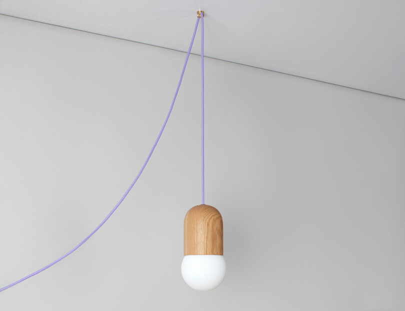 A modern pendant light from the Light Bean collection by Katerina Kopytina features a wooden top and white bulb, suspended by a purple cord against a plain light gray background.