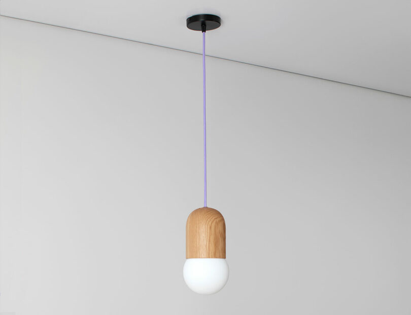 A modern pendant light from the Light Bean collection by Katerina Kopytina features a wooden top and white bulb, suspended by a purple cord against a plain light gray background.