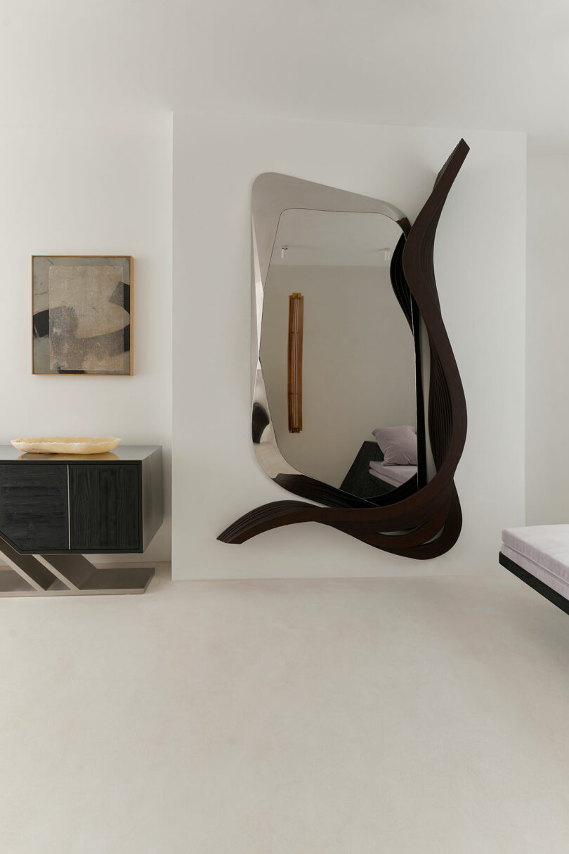 A modern room with a large abstract Miminat mirror, a geometric sideboard, an abstract wall art piece, and part of a bed visible on the right.