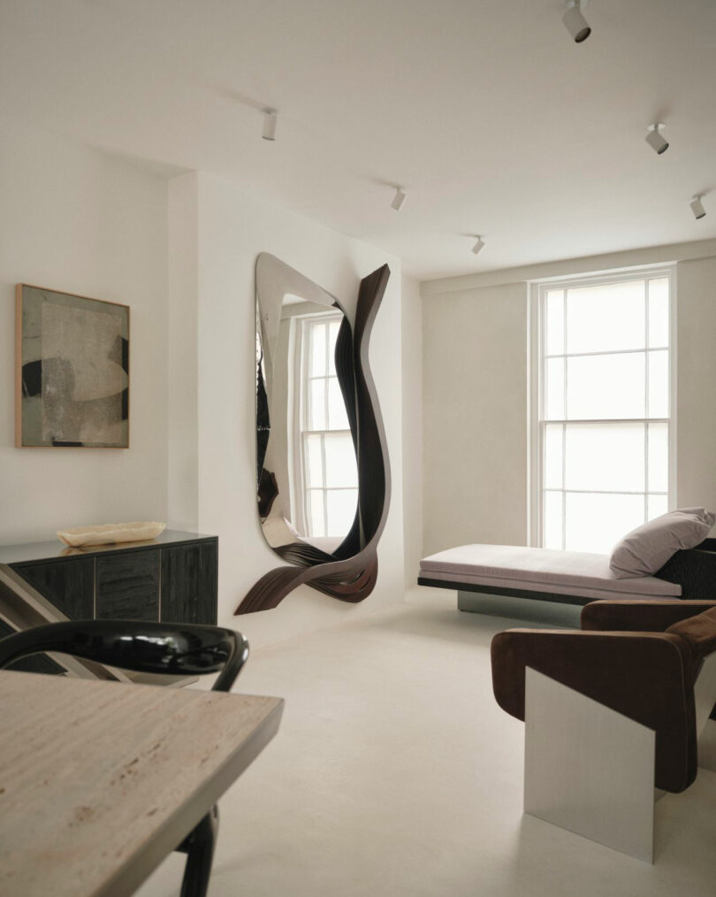 A modern Miminat minimalist room with neutral tones features a large sculptural wall mirror, a daybed by tall windows, abstract art, and sleek contemporary furniture.