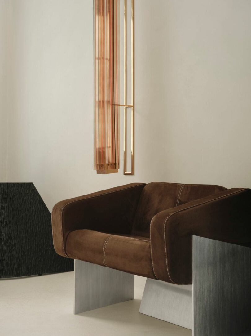 A modern brown suede Miminat armchair with metal legs sits beside a black textured side table and a vertical gold and glass pendant light against a neutral wall.
