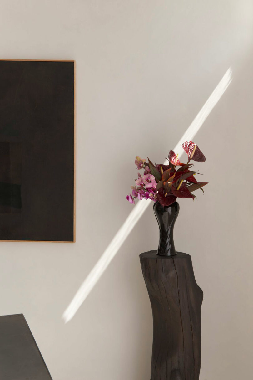 A black vase with assorted flowers, from Miminat, sits on a wooden pedestal beside a dark rectangular wall art; a beam of sunlight cuts diagonally across the pale wall.