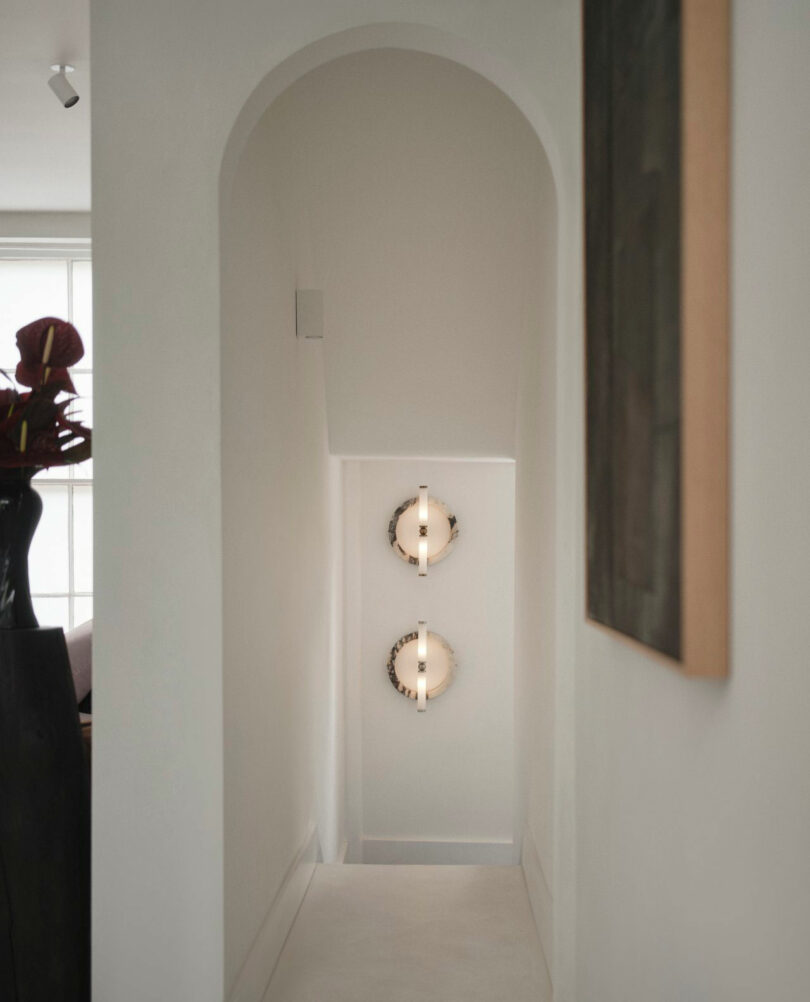 A narrow Miminat hallway with an arched ceiling features two round wall lights and minimal decor, with light streaming in from an adjacent room.