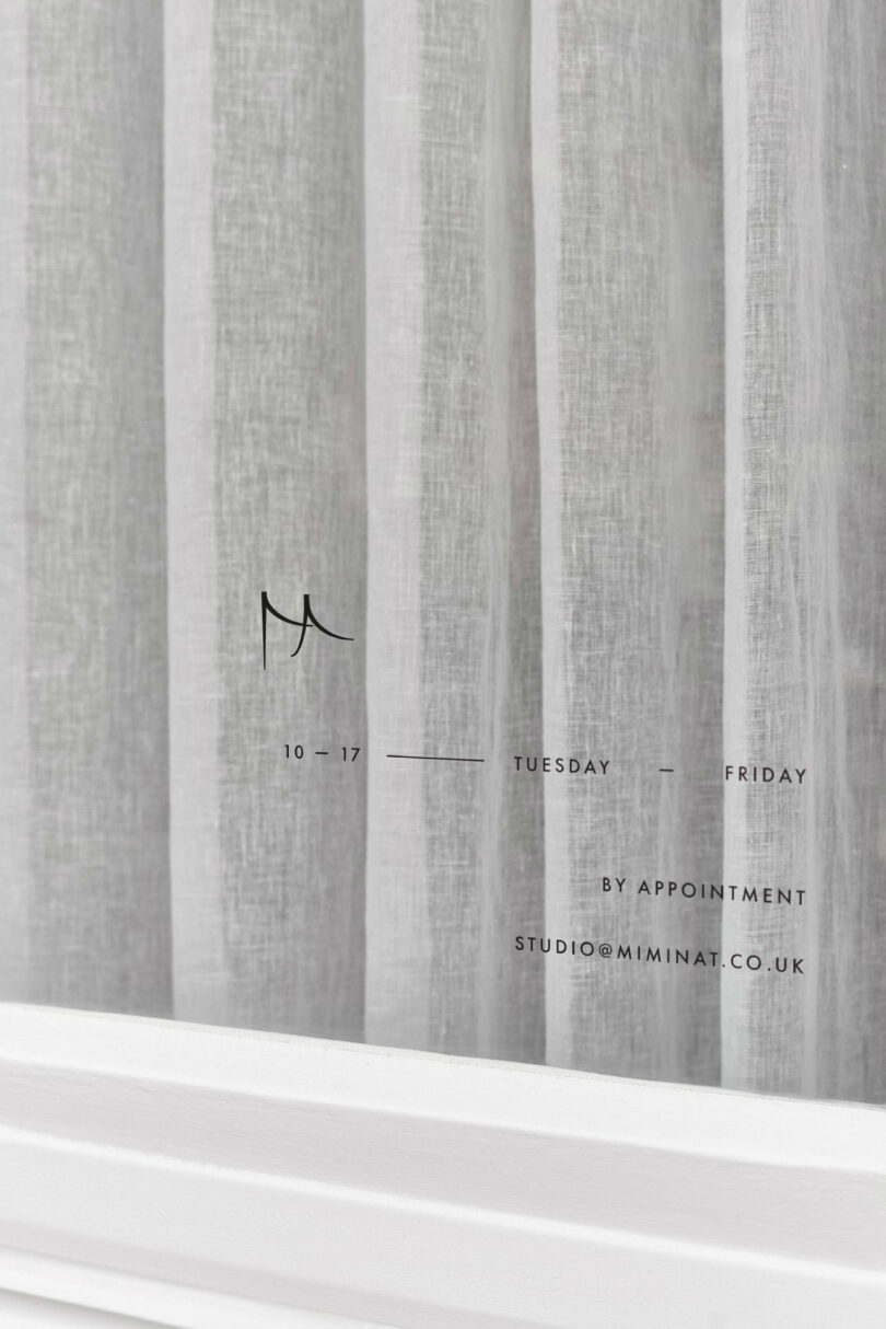 White sheer curtains with vertical folds frame the window. Minimalist black text displays Miminat’s business hours, appointment info, and an email address for studio@miminat.co.uk.
