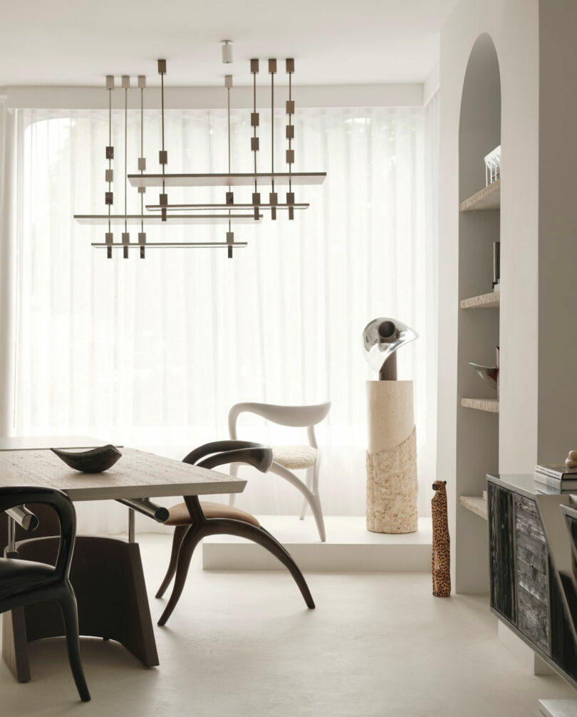 Minimalist living space with sculptural chairs, a geometric chandelier, abstract decor on a pedestal, built-in shelves, and sheer curtains letting in natural light—all contributing to a refined Miminat-inspired design.