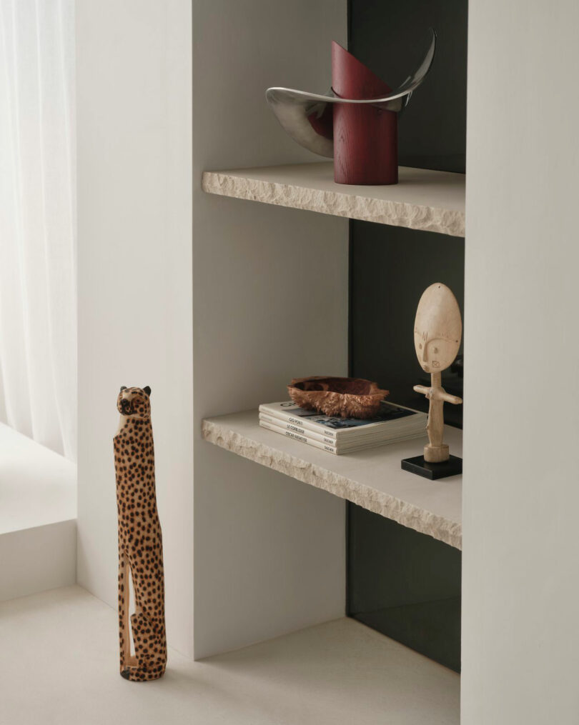Miminat-inspired minimalist shelves display decorative objects, including a red and silver sculpture, books, a carved wooden figure, a small tray, and a tall ceramic cheetah statue.
