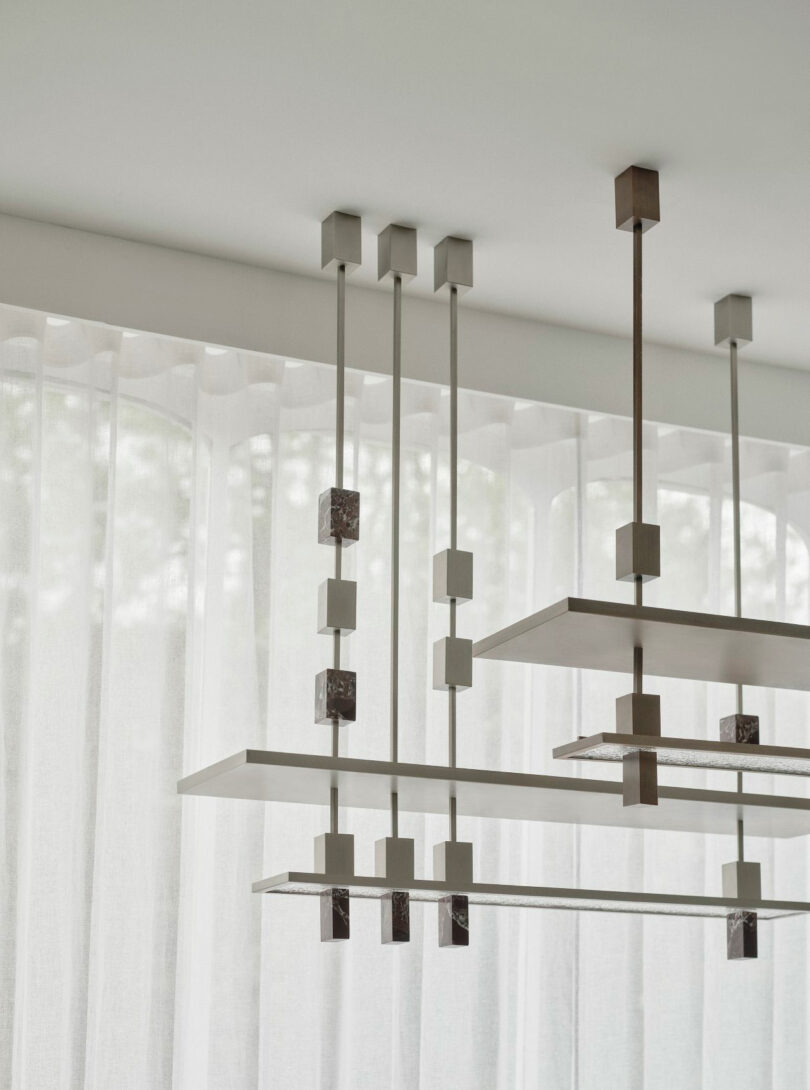 Modern geometric light fixtures by Miminat with rectangular shelves and cube accents hang from a white ceiling in front of sheer white curtains.