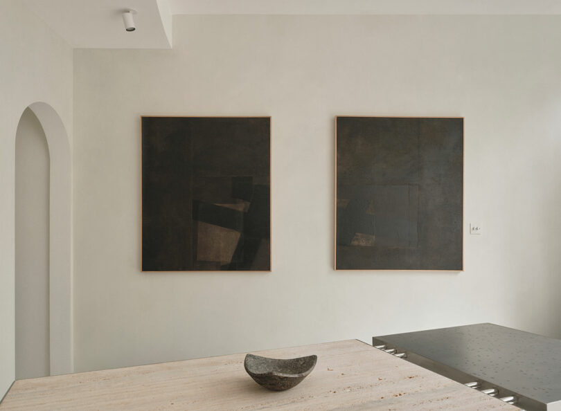 Minimalist interior with two dark abstract paintings on a light wall, a recessed ceiling light, an arched doorway, and a stone bowl on a light-colored table embody the essence of Miminat design.
