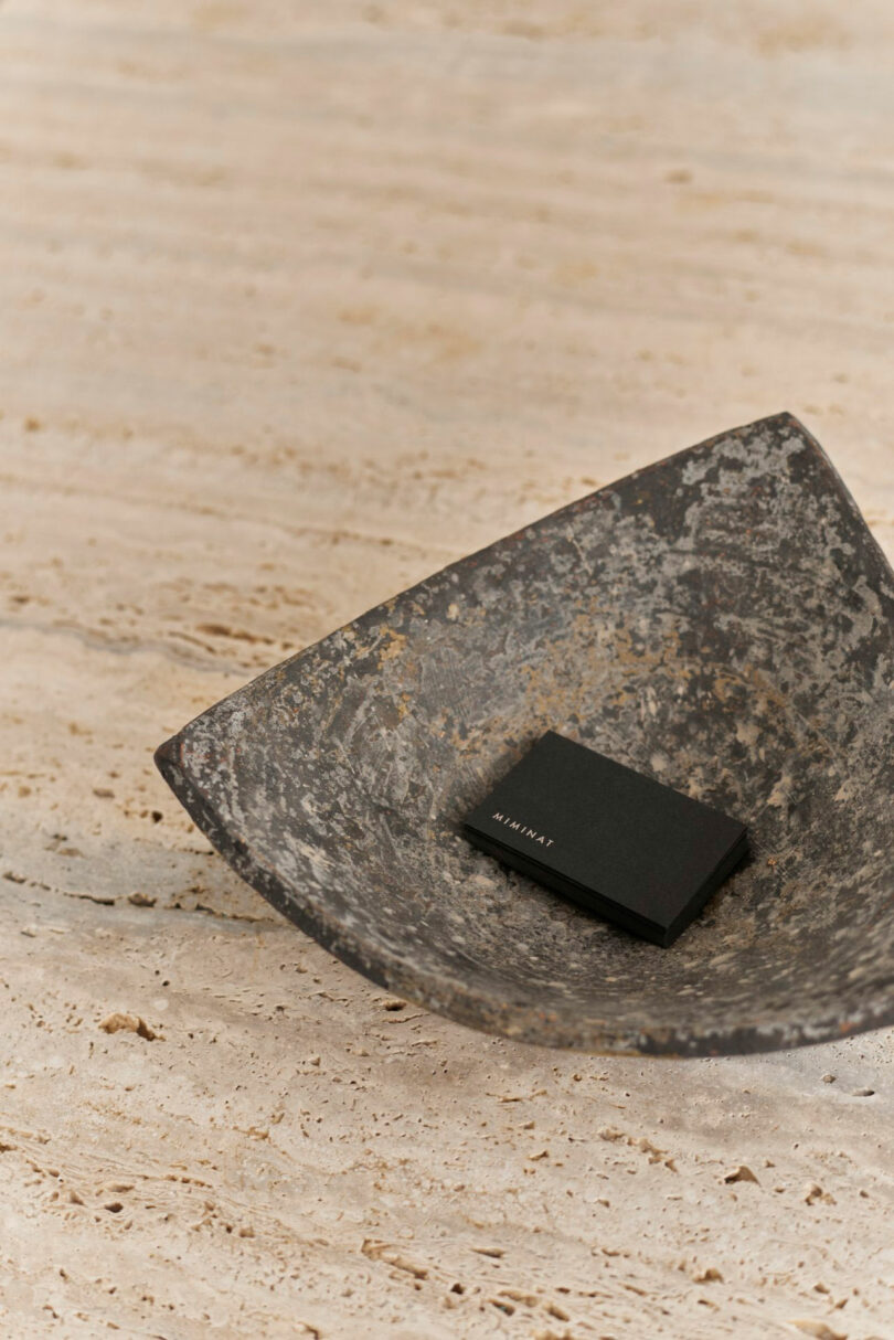A black rectangular Miminat object rests in a weathered, shallow metal bowl placed on a beige stone surface.