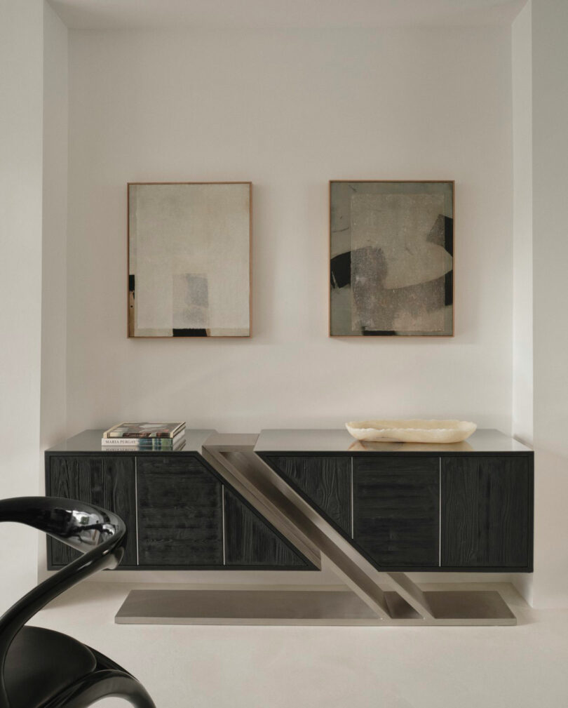 Minimalist interior with a Miminat black console table, sculptural metal accent, stacked books, a shallow bowl, and two abstract framed artworks on a white wall.