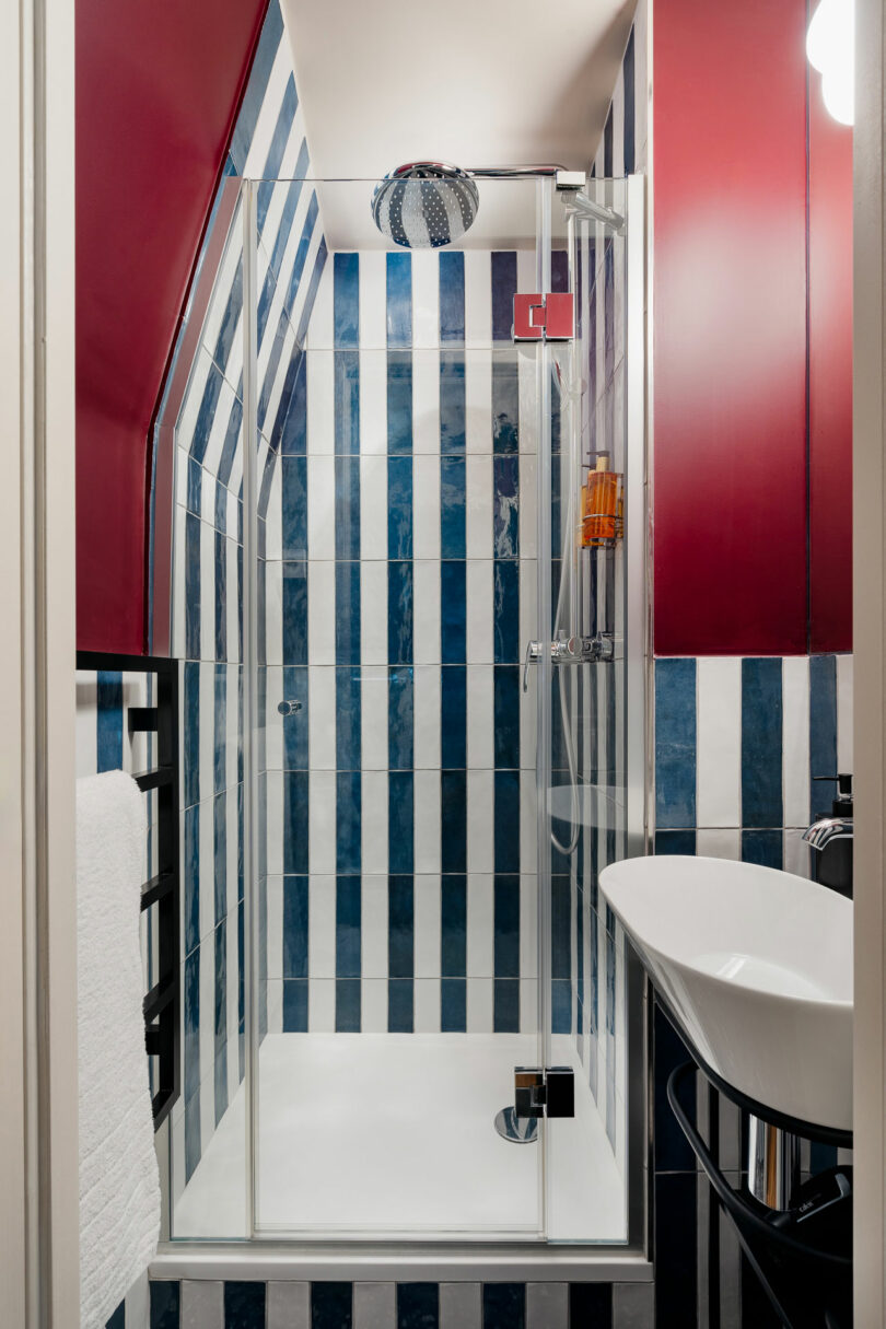 A compact bathroom with a glass shower featuring vertical blue and white striped tiles, a white basin sink, red cabinets, and a towel rack.