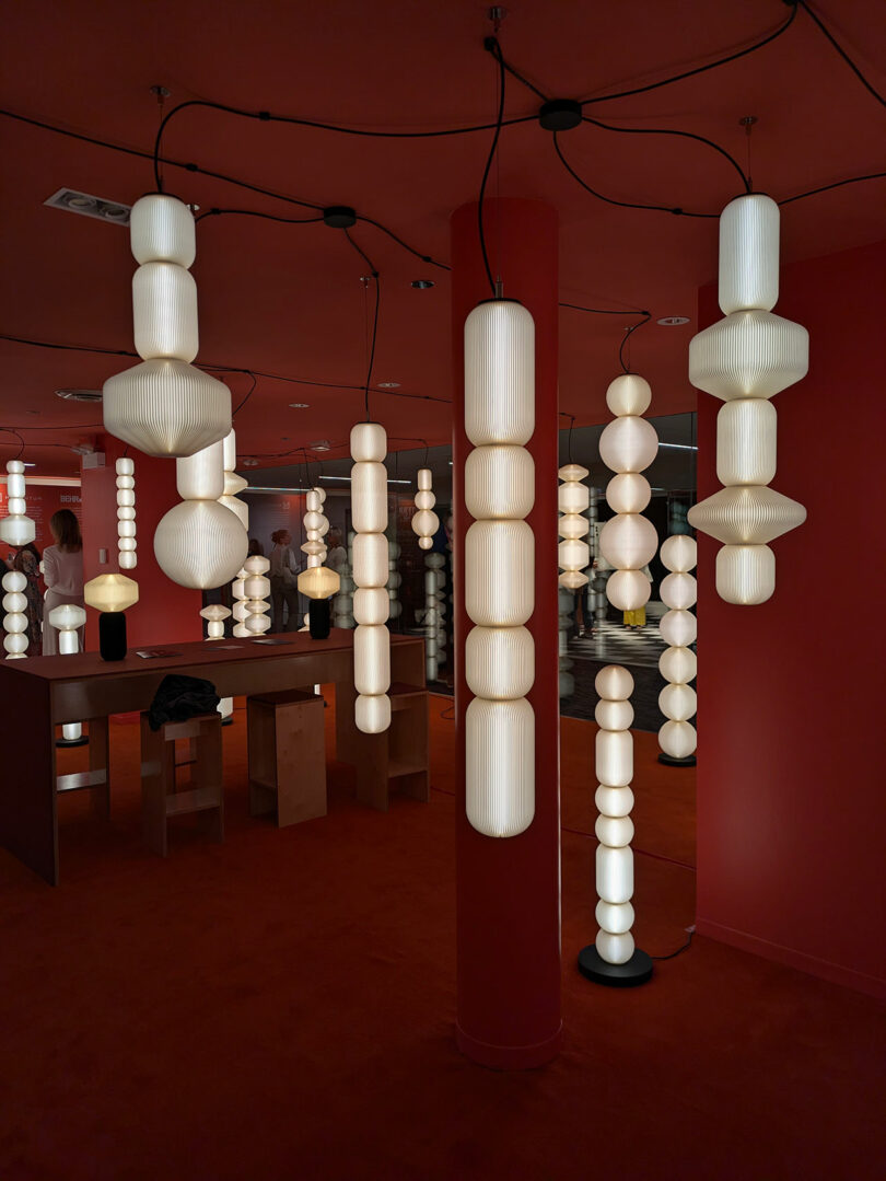 Hanging cylindrical and geometric white lights illuminate a red room with tables, stools, and a central pillar—a striking space worthy of NeoCon 2025 Top 10 distinction.