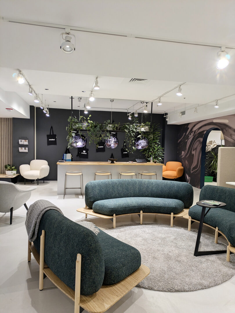 Modern office lounge with green curved sofas, a round gray rug, chairs, a counter with stools, wall-mounted screens, and plants hanging above the work area—inspired by NeoCon 2025 Top 10 design trends.