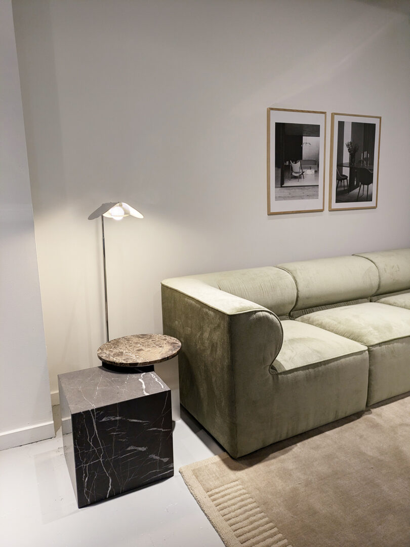 A modern living room with a green velvet sofa, a marble side table, a lamp, a beige rug, and two framed black and white photos on a white wall.
