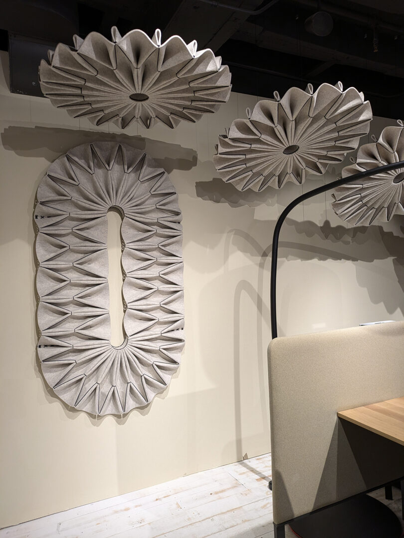 Wall-mounted and ceiling acoustic panels with a pleated, fabric design are displayed above and beside a beige booth and wooden table in a modern interior space.