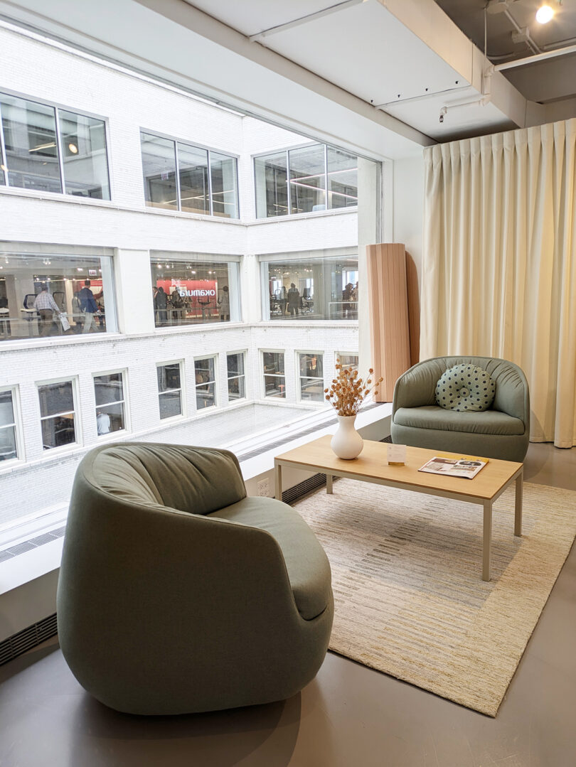 Two sage green armchairs and a light wood coffee table on a beige rug by a large window in a modern, bright office lounge area.