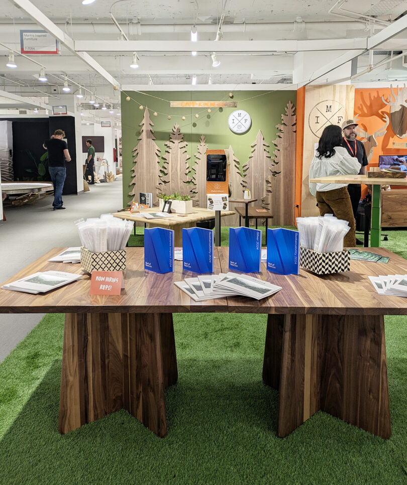 A display table with brochures and plastic-wrapped items stands on artificial grass in front of a booth with a forest-themed backdrop and wooden decorations at an indoor event.