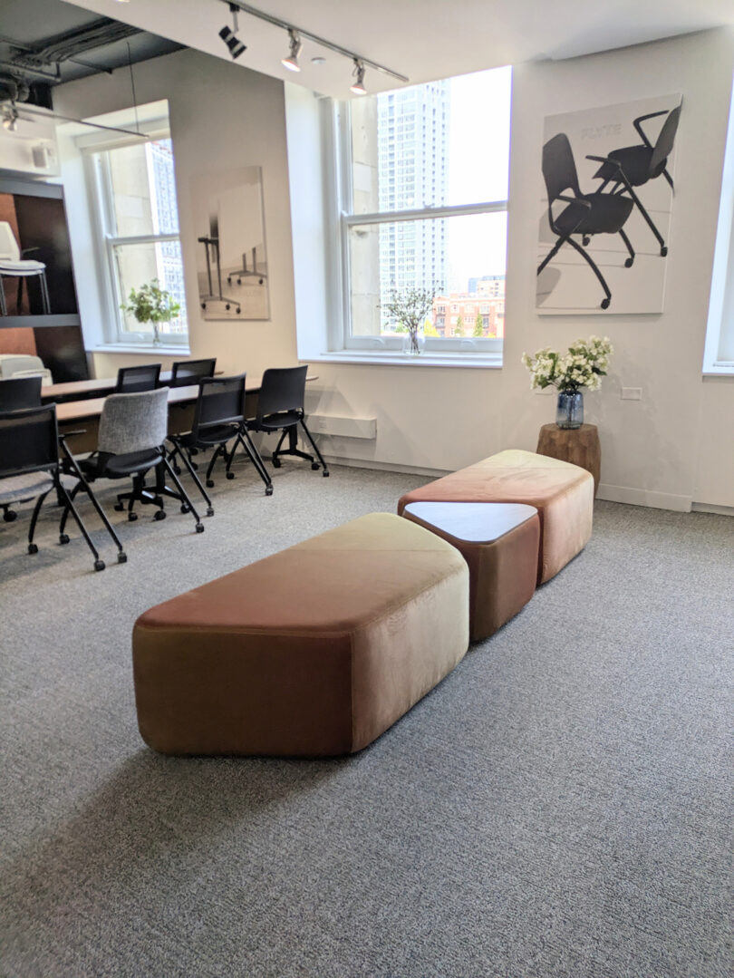 Modern office space with a meeting table and chairs, large windows, abstract art on walls, and cushioned benches in the foreground.