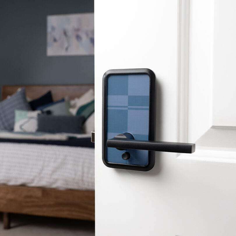 A blue checkered modern smart door lock is installed on a white door, partially open to reveal a blurred bedroom interior.