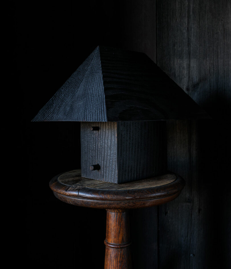 A dark wooden birdhouse from the Ravine Collection Post Company, with a triangular roof, sits on a round wooden stool against a dark background.