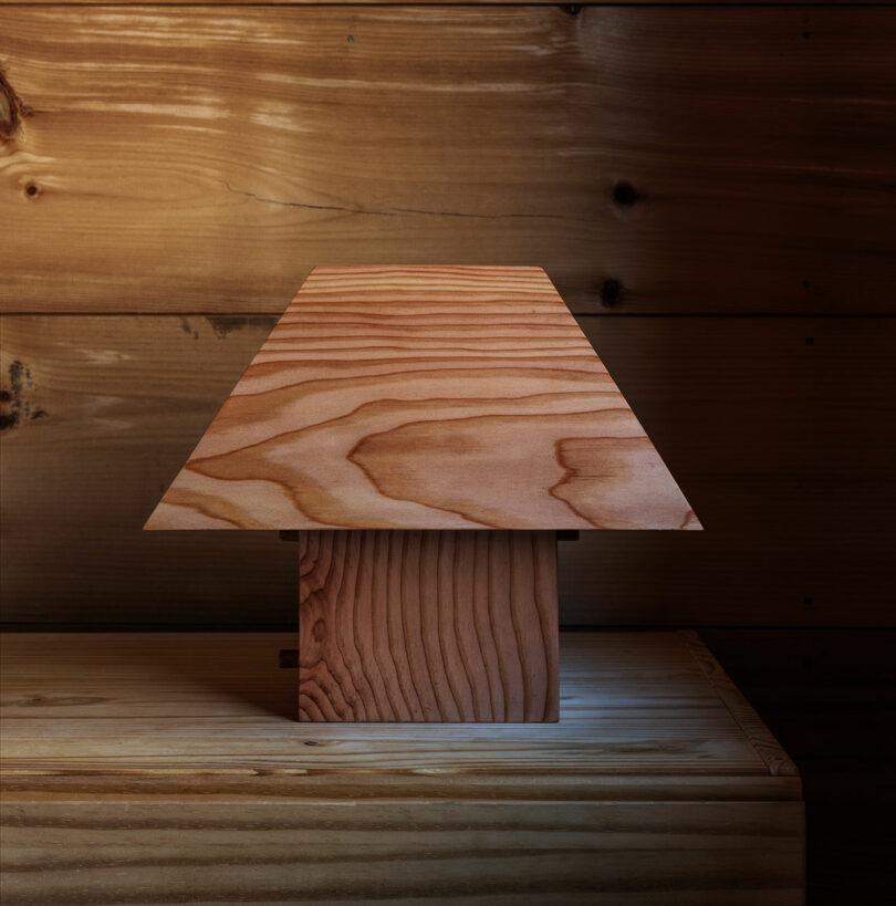 A wooden lamp from the Ravine Collection Post Company, featuring a square base and wide, flat shade, sits on a wooden surface against a wooden plank wall.