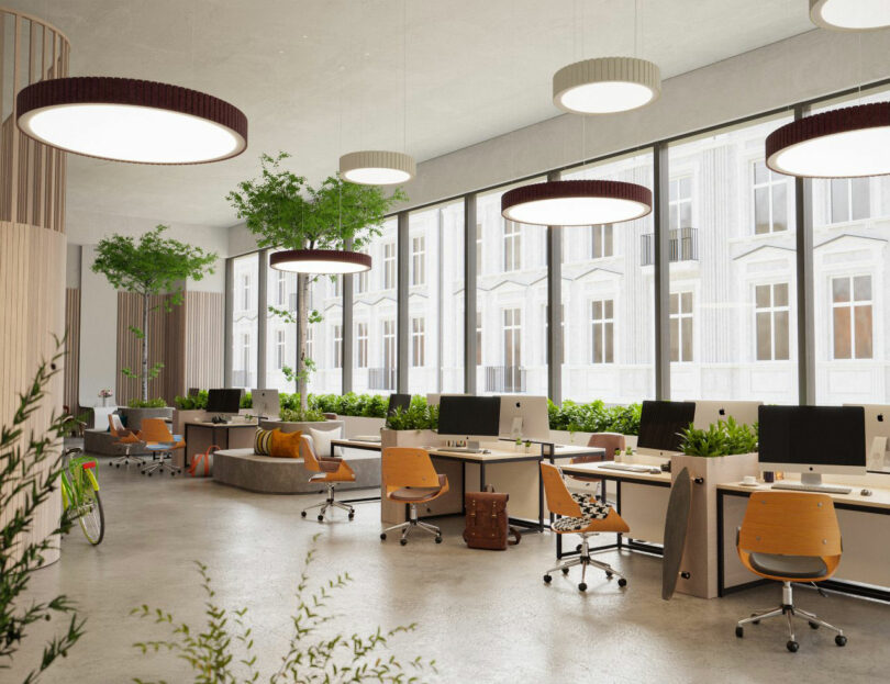 A modern open-plan office with large windows, wooden desks, computer monitors, orange chairs, indoor plants, and circular ceiling lights.