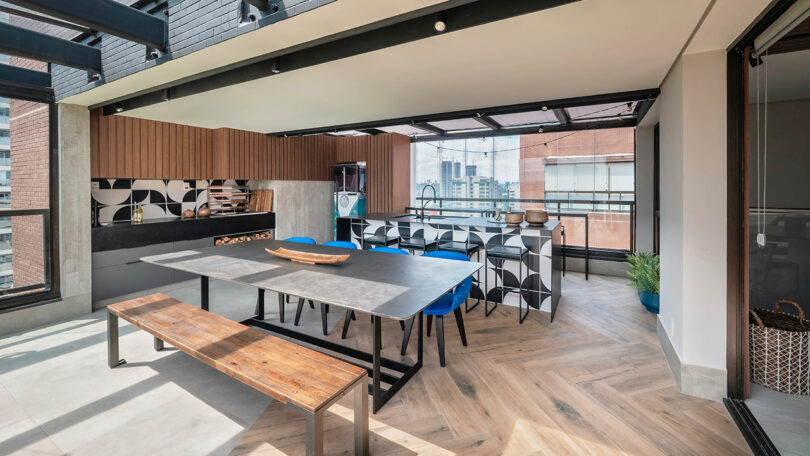 Modern covered outdoor kitchen and dining area with a long table, wooden bench, grill, patterned tiles, blue chairs, and city view through glass railing.