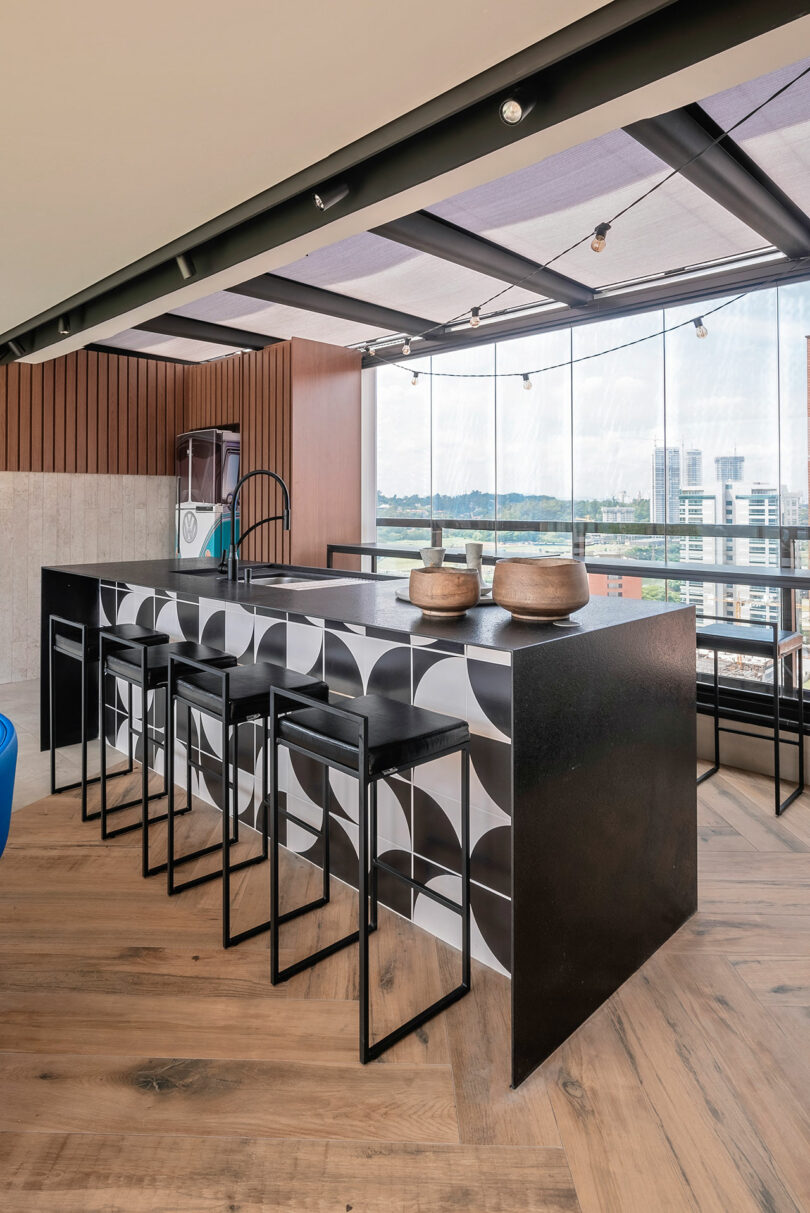 Modern kitchen with black and white geometric island, four black stools, wooden bowls on the counter, wood flooring, and large windows overlooking a cityscape.