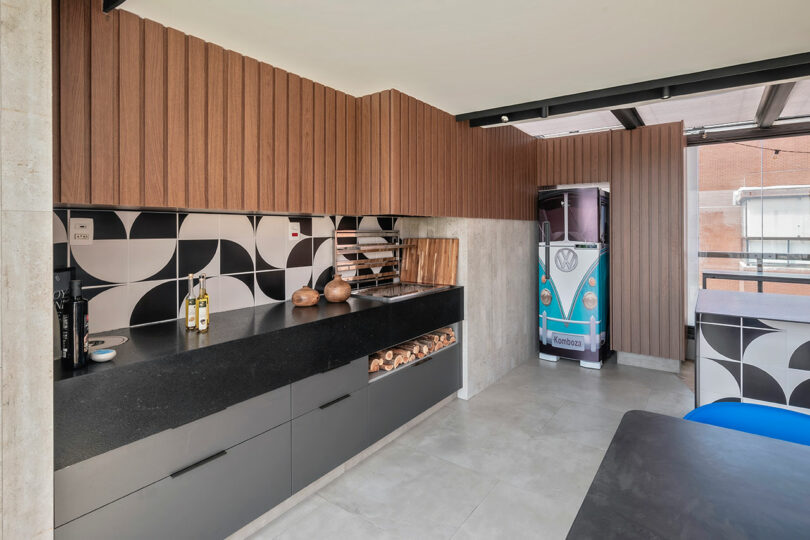 Modern kitchen with geometric black-and-white backsplash, wooden cabinets, black countertop, open shelves with firewood, and a blue retro-style refrigerator shaped like a van.