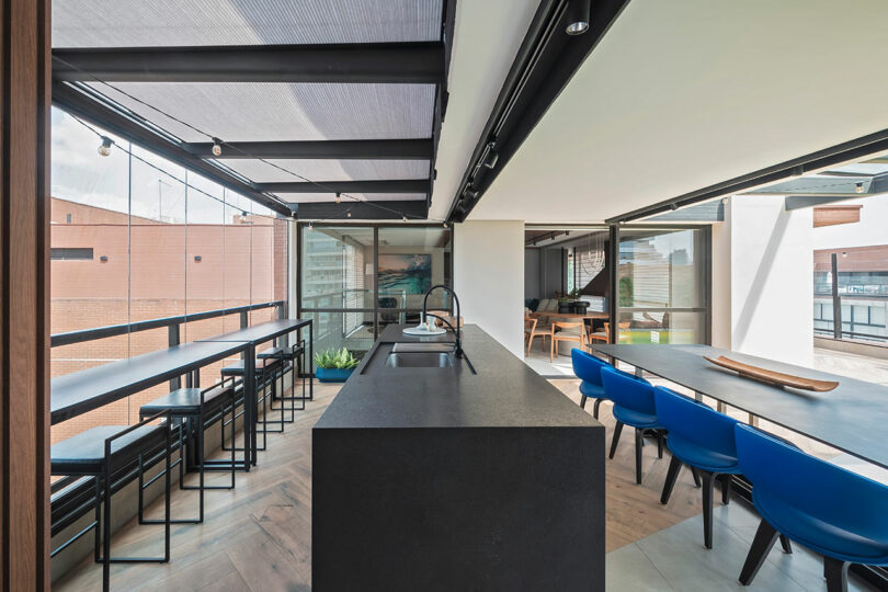 Modern open-plan dining and kitchen area with a black island, high bar seating by large windows, and a long table with blue chairs. Natural light fills the space.