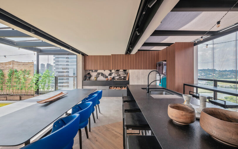 Modern open kitchen and dining area with black countertops, blue chairs, wooden accents, and large windows overlooking a cityscape and greenery.
